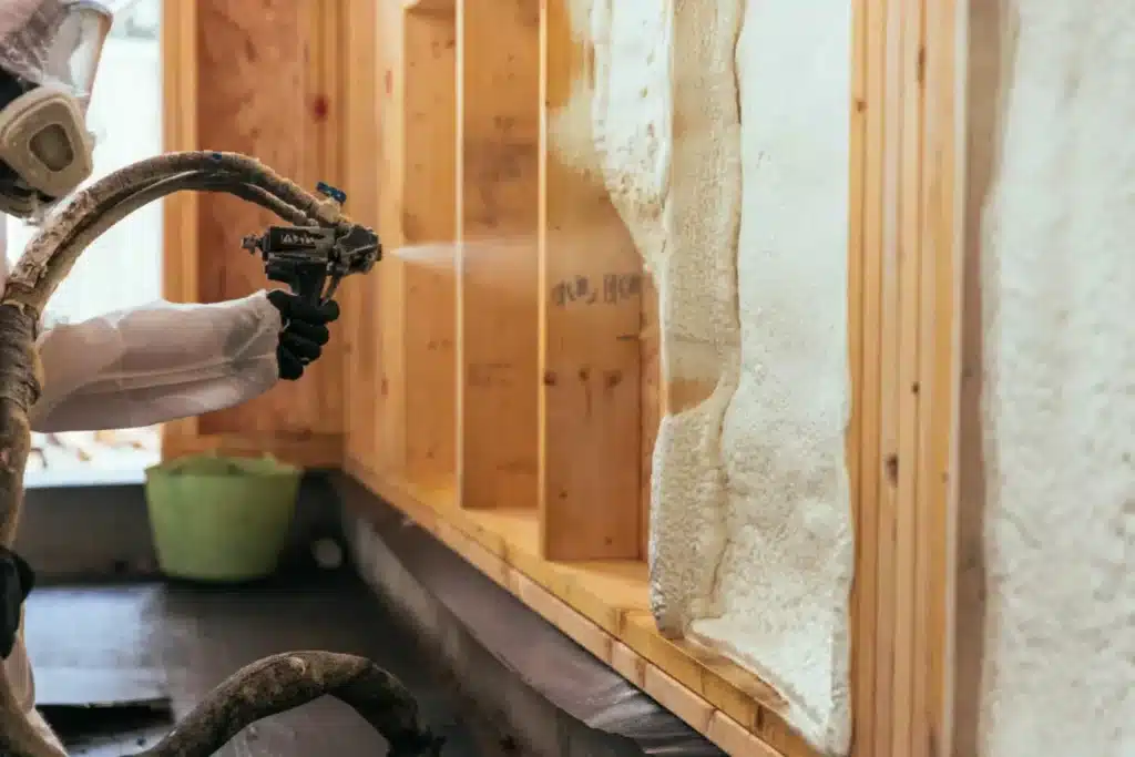 Worker in full protective gear, including a respirator, spraying white foam insulation into the lower cavities of a wood-framed wall.